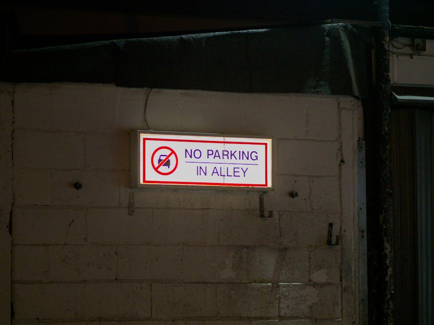 A circular no parking road sign with a blue background and a red border, mounted on a metal post standing on a paved area in front of an old brick wall with cream, beige, and dark stones. The sign casts a shadow on the wall due to sunlight. The brick wall has a horizontal red brick line near the bottom, with weathered bricks and mortar visible. The scene is outdoors, likely in a residential or urban setting, and is relevant to house removals and parking regulations for moving services, with the sign indicating restrictions on parking or loading in the area. Man with Van New Malden operates in this location, providing professional moving and furniture transport services, which may involve loading and unloading within such parking restrictions during home relocation or house clearance processes.