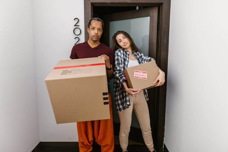 A man and a woman stand inside a doorway, each holding a cardboard box as part of a home relocation or furniture transport process. The man, positioned on the left, is wearing a maroon t-shirt and orange trousers, holding a large cardboard box with black tape along the edges and a shipping label. His facial expression is neutral, and he is looking directly at the camera. The woman, on the right, is dressed in a plaid shirt over a white top and beige pants, holding a smaller rectangular cardboard box with a red 'Fragile' sticker on it. She is leaning her head slightly to the side with a relaxed expression. Behind them, the door frame is dark wood, and the door is open. The space appears to be the entrance or hallway of a property, with light-colored walls and minimal decoration. The scene is lit with natural or soft indoor lighting, capturing a moment in the packing and loading process for a professional removals service provided by Man with Van New Malden, with a focus on careful handling of household items during a house move.