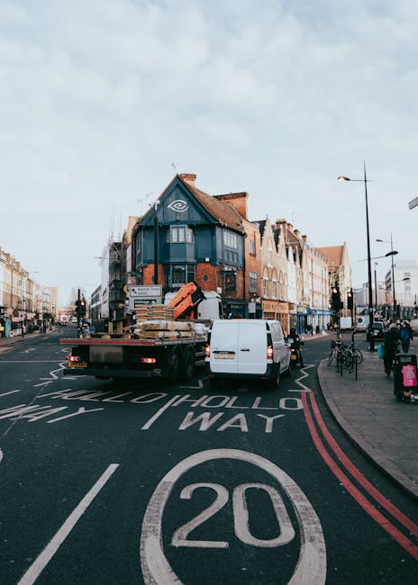 A flatbed truck belonging to Man with Van New Malden is parked in the middle of New Malden High Street during daylight hours, with the truck loaded with multiple wooden pallets and cardboard boxes secured with straps and plastic wrap, indicating preparations for a home relocation or furniture transport. The truck is positioned near a crosswalk, with a white van parked directly behind it. The scene includes a row of commercial premises with brick facades and large windows on either side of the street, and pedestrians walking along the pavement carrying shopping bags and backpacks. Street lamps and bicycles are visible along the sidewalk, and the sky is partly cloudy. The loading process appears to be ongoing, with items awaiting transport into a nearby property. Natural lighting highlights the urban environment, reflecting typical house removals and moving logistics in the New Malden area.