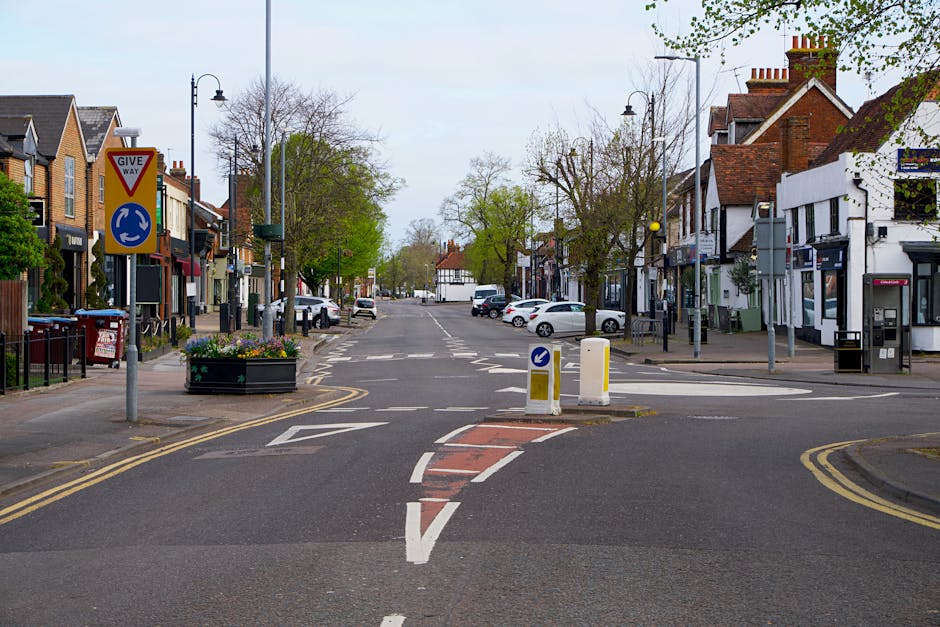 A wide view of New Malden High Street showing a central roundabout with traffic bollards and directional signage, including a 'give way' sign. On both sides of the street, there are shops, cafes, and residential buildings with brick exteriors and pitched roofs. Trees with early spring foliage line the pavement, and parked cars are visible along the road. Street lamps and flower planters add to the street’s aesthetic. The scene appears to be during daytime with overcast skies, providing natural lighting for the area. This setting reflects an urban environment suitable for house removals or furniture transport, where the loading process might involve navigating through commercial and residential areas, supported by companies like Man with Van New Malden for efficient home relocation services.