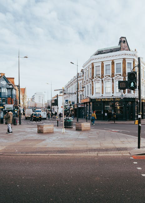 A flatbed truck belonging to Man with Van New Malden is parked in the middle of New Malden High Street during daylight hours, with the truck loaded with multiple wooden pallets and cardboard boxes secured with straps and plastic wrap, indicating preparations for a home relocation or furniture transport. The truck is positioned near a crosswalk, with a white van parked directly behind it. The scene includes a row of commercial premises with brick facades and large windows on either side of the street, and pedestrians walking along the pavement carrying shopping bags and backpacks. Street lamps and bicycles are visible along the sidewalk, and the sky is partly cloudy. The loading process appears to be ongoing, with items awaiting transport into a nearby property. Natural lighting highlights the urban environment, reflecting typical house removals and moving logistics in the New Malden area.