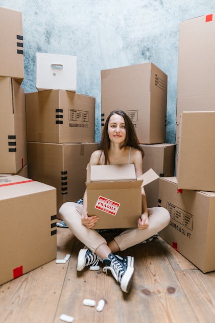 A young woman with long brown hair, dressed in casual beige clothing and sneakers, sitting cross-legged on a wooden floor inside a room surrounded by stacked cardboard boxes of various sizes, some labeled with red 'fragile' stickers. She holds an open cardboard box on her lap, appearing to prepare for a home relocation or furniture transport. The boxes are sealed with packing tape and some have printed labels, indicating packing and moving preparations. The background features a textured light blue wall, and the scene is lit with natural or soft artificial lighting, reflecting a typical environment for professional removals or packing services carried out by companies like Man with Van New Malden during a furniture transport or moving process.