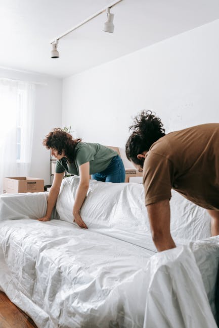 In a bright, minimally decorated room with white walls and natural daylight coming through a window, two movers from Man with Van New Malden are in the process of preparing furniture for home relocation. One person, dressed in a green shirt, is standing on a sofa and smoothing out a white protective cover made of fabric or plastic wrap over the cushions. The other individual, wearing a brown shirt, is partially visible and appears to be securely fastening or adjusting the covering. The sofa is positioned against the wall, with cardboard boxes and packing materials visible on a side table and in the background, indicating an ongoing packing or furniture transport process. The image captures a calm moment of packing and furniture protection, essential steps in house removals, especially for navigating tight stairs or narrow doorways typically involved in residential moves.