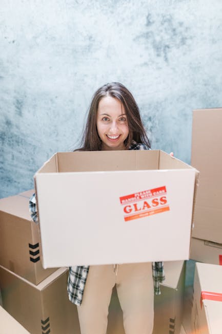 A young woman with long brown hair, dressed in casual beige clothing and sneakers, sitting cross-legged on a wooden floor inside a room surrounded by stacked cardboard boxes of various sizes, some labeled with red 'fragile' stickers. She holds an open cardboard box on her lap, appearing to prepare for a home relocation or furniture transport. The boxes are sealed with packing tape and some have printed labels, indicating packing and moving preparations. The background features a textured light blue wall, and the scene is lit with natural or soft artificial lighting, reflecting a typical environment for professional removals or packing services carried out by companies like Man with Van New Malden during a furniture transport or moving process.