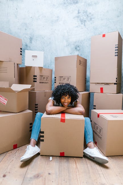 A young woman with curly black hair, wearing a white t-shirt, blue jeans, and white sneakers, sitting on a light wooden floor surrounded by numerous cardboard boxes of varying sizes, some sealed with