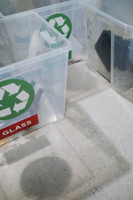 A close-up view of two clear plastic recycling bins with green recycling symbols on their sides, placed on a light-colored carpeted floor inside a property. One of the bins has a red label marked 'GLASS,' indicating its designated use for glass waste. The bins are positioned near a doorway or entrance, with a shadow of the photographer visible on the carpet. The background shows part of a wall and a section of another bin or container. This image captures the process of sorting and preparing recyclable materials as part of a home relocation or moving service, illustrating the organization required during packing and waste disposal handled by Man with Van New Malden, a professional removal company specializing in house removals and cleaning up after packing.