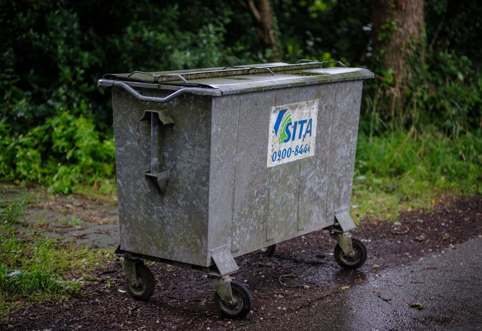 A large, metal wheelie bin with a grey, weathered surface sits on an outdoor pavement area adjacent to a grassy verge with shrubbery and trees in the background. The bin features a closed, dark lid and sturdy black wheels, with a blue and white SITA logo and customer service number visible on its front. The surrounding environment appears damp, with patches of wetness on the ground, indicating recent rain. The scene is well-lit with natural daylight, showcasing the bin ready for collection as part of home relocation or waste disposal services, consistent with the focus of a professional removals process via Man with Van New Malden. This setting captures the typical outdoor scene where waste bins are placed for collection in the context of home or garden clearance during moving or disposal activities.