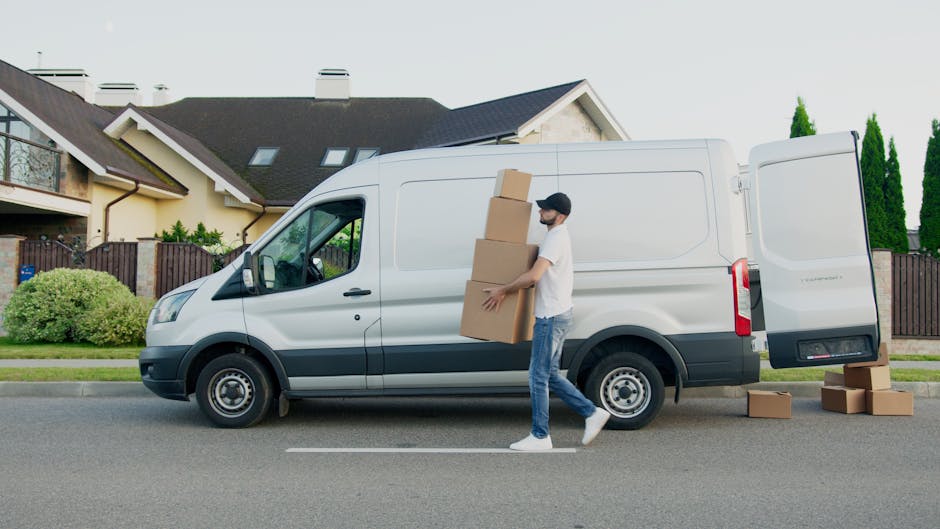 A young man with a beard, wearing a red cap, a checkered shirt, and a two-tone brown and beige jacket, stands inside the rear of a van parked on a driveway. The van’s interior is visible, with sever