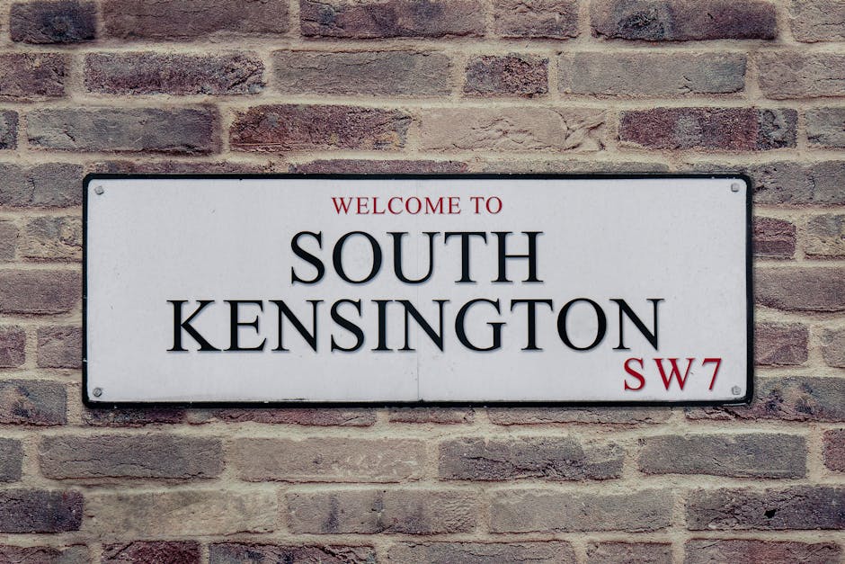 Photograph of a rectangular white metal street sign mounted on a brick wall. The sign displays the words 'Welcome to South Kensington' in bold black letters, with 'South Kensington' in larger font, and 'SW7' in smaller red text at the bottom right corner. The brick wall behind the sign consists of reddish-brown bricks arranged in a traditional pattern, with some variation in color and texture, and is illuminated by natural daylight. The sign is securely attached with visible screws at each corner. The image reflects a typical residential or commercial building exterior in London, an area associated with house removals and relocations, supporting the context of moving services like those offered by Man with Van New Malden.