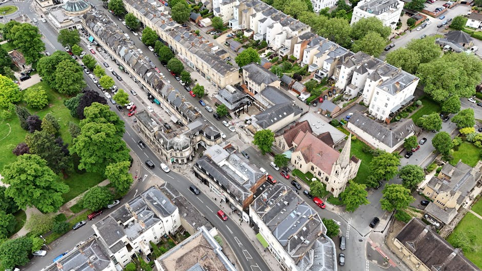 An aerial view of a residential area in New Malden showing a mix of terraced houses, semi-detached properties, and a small church with a tall steeple. The scene features a narrow, winding street lined with parked cars, including small hatchbacks and sedans, with some vehicles moving slowly along the road. The street is intersected by side roads, with trees and green spaces situated between the houses, providing lush foliage and shade. Visible within an open driveway area is a van being loaded or unloaded with various packed boxes and furniture, wrapped in plastic and protective materials, indicative of a home relocation process. A trolley and moving blankets are nearby, supporting the logistics of furniture transport. The overall scene reflects a typical neighbourhood prepared for a house removal or moving service, with the presence of a van from Man with Van New Malden hinting at local removals activities, possibly involving packing, loading, and transporting household items during a house move.