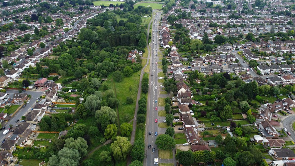 An aerial view of a residential area in New Malden, showing a street lined with terraced houses, some with pitched roofs and chimneys, and a mix of trees and greenery along the sidewalks. A church wit