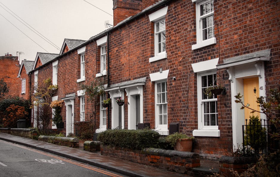 Photograph of a row of traditional red brick terraced houses on a narrow street, showcasing white-framed sash windows and doorways, with some houses having small window awnings and potted plants near 