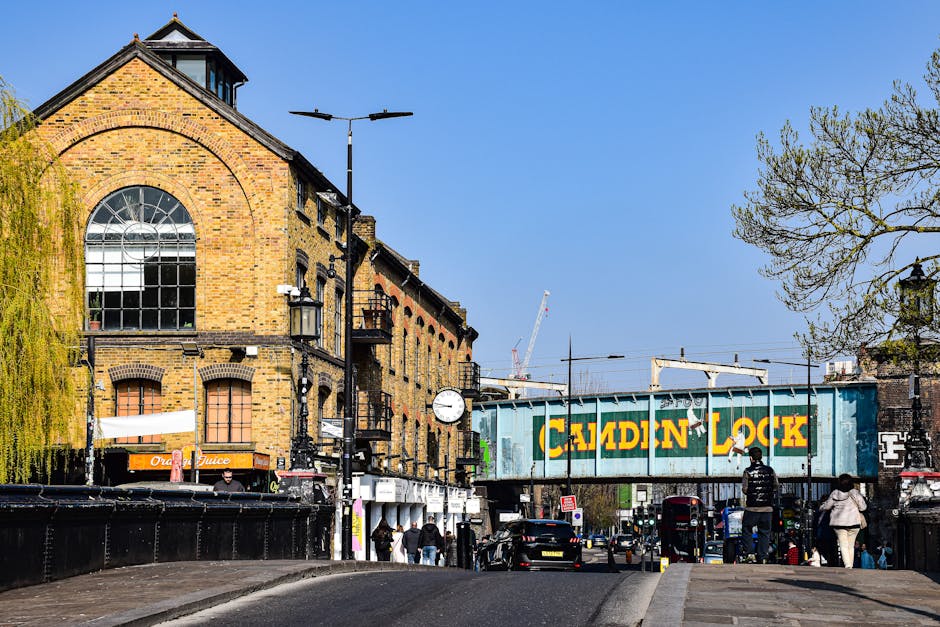 A street scene in New Malden featuring a large brick building with a prominent arched window, situated next to a cast iron clock mounted on a lamppost. To the right, a metal railway bridge with the words 'CALEDIN LOCK' painted in yellow spans across the street, indicating an active railway line. In the foreground, several pedestrians are walking on the pavement, some nearby parked cars, while a man is seen loading or unloading items from a small moving van or vehicle, which is partially visible at the edge of the image. The area includes various shopfronts, including a juice bar, with urban trees on the right and a clear blue sky overhead. The scene suggests activities related to packing, loading, and home relocation, aligned with house removals and furniture transport services, such as those offered by Man with Van New Malden.